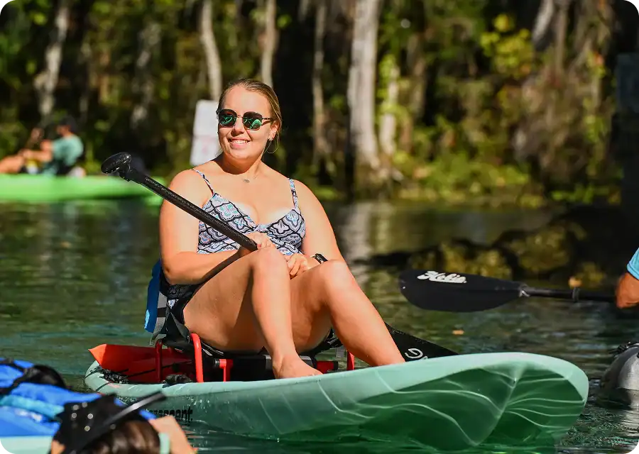 A smiling woman in sunglasses sitting in a light green kayak on a sunny river